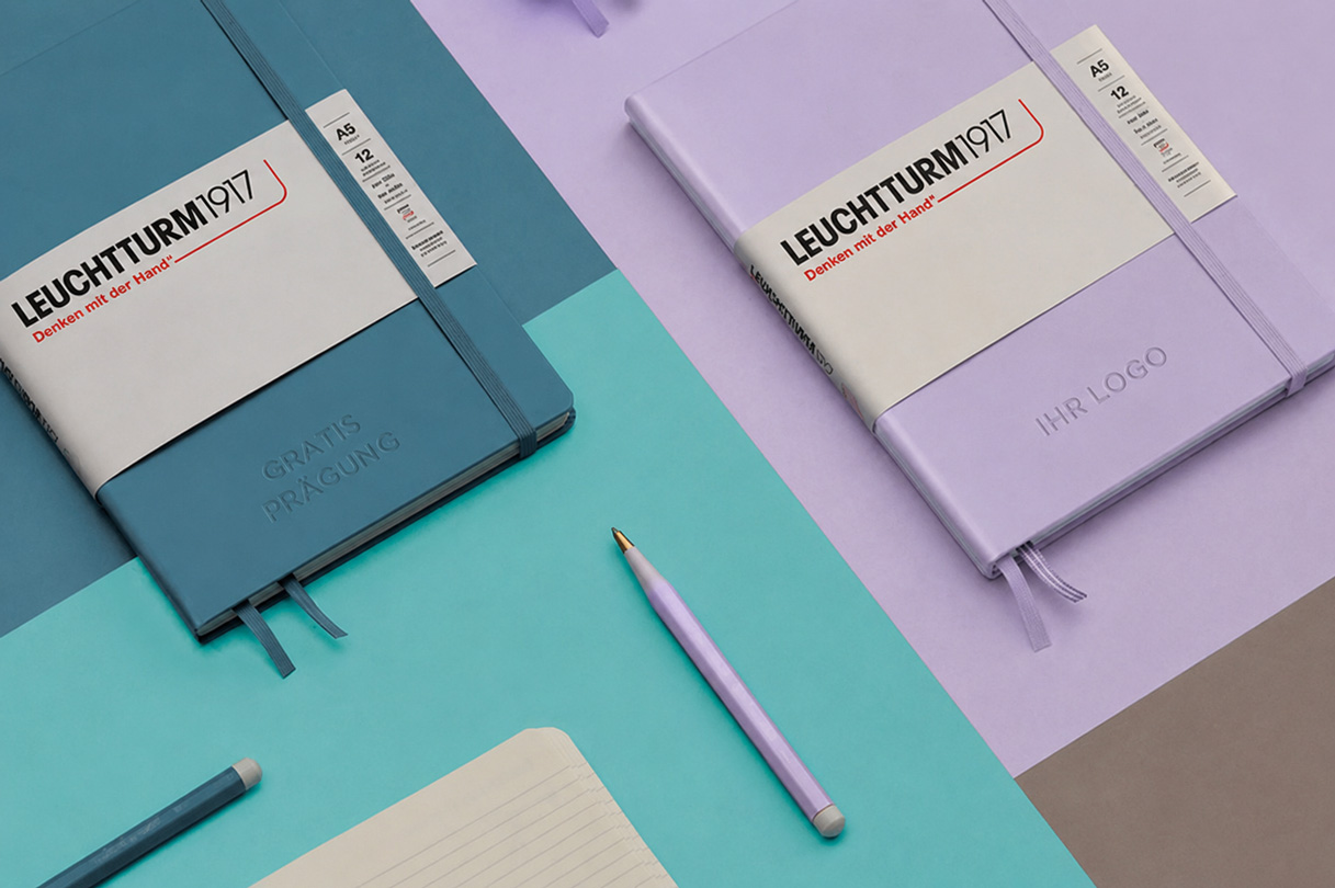 Zwei Leuchtturm1917 Notizbücher in trendigen Farbtönen mit gratis Blindprägung und individuellem Logo auf modernem Hintergrund inszeniert.